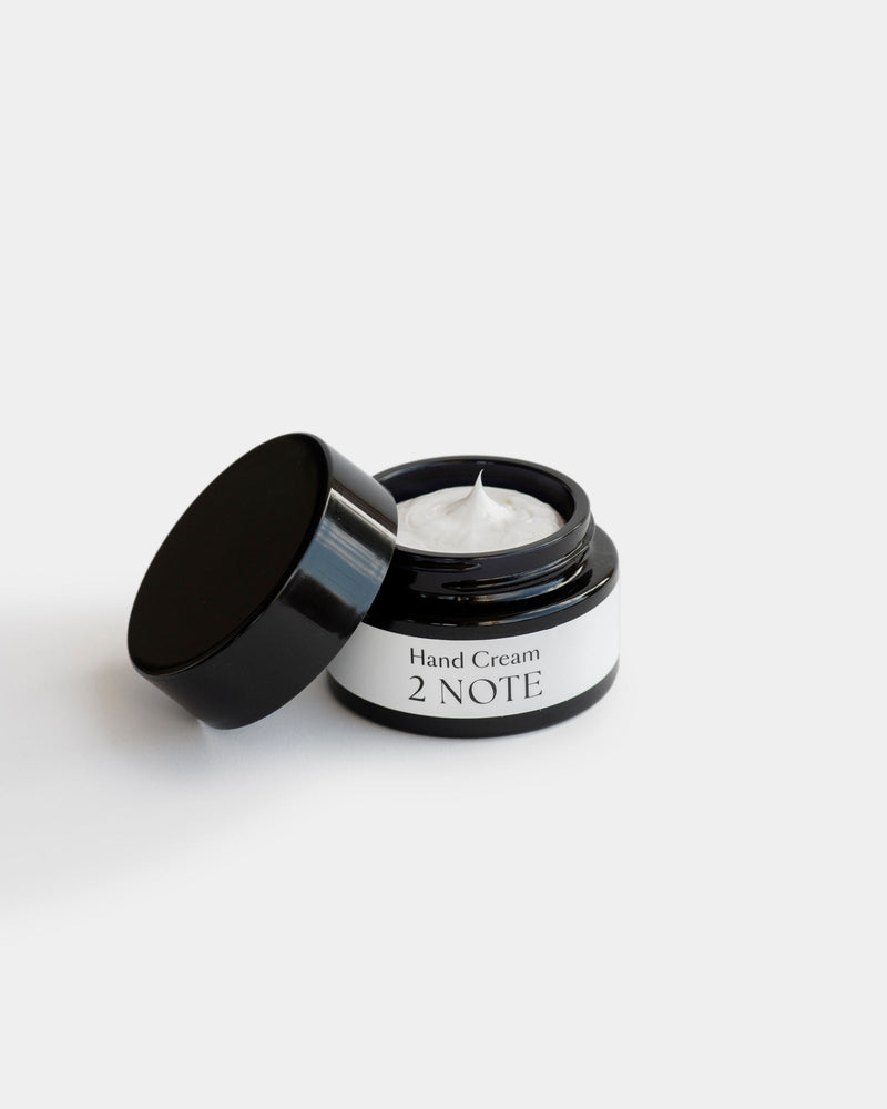 Hand Cream in open matte black glass jar, lid leaning beside it — natural skincare cream texture visible inside, light gray background