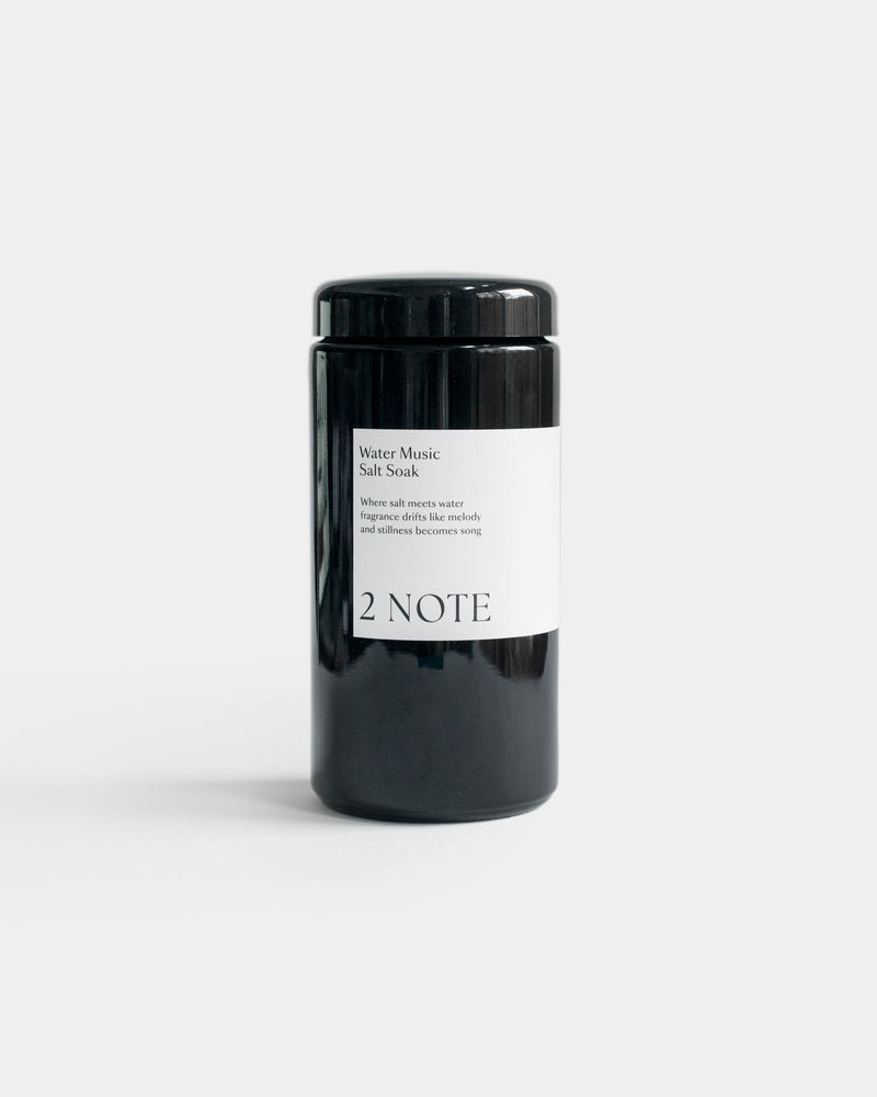 Water Music Salt Soak in tall matte black cylindrical jar — botanical natural skincare bath soak with essential oils, front view on light gray