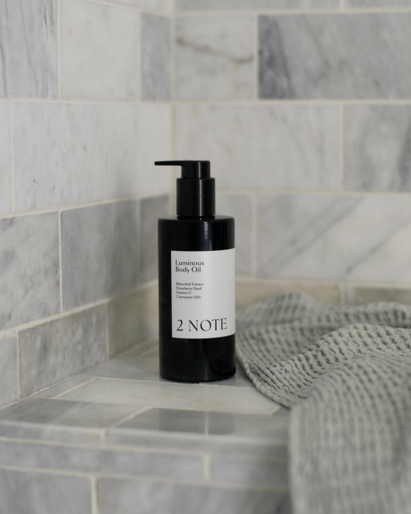 Luminous Body Oil matte black pump bottle on marble tile shower shelf beside gray waffle towel — botanical skincare brightening body oil in bathroom setting