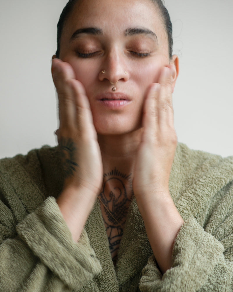 Queer nonbinary model with light olive complexion and curly brown hair in a bun, eyes closed, pressing palms to face — applying Cleansing Face Oil, natural skincare application