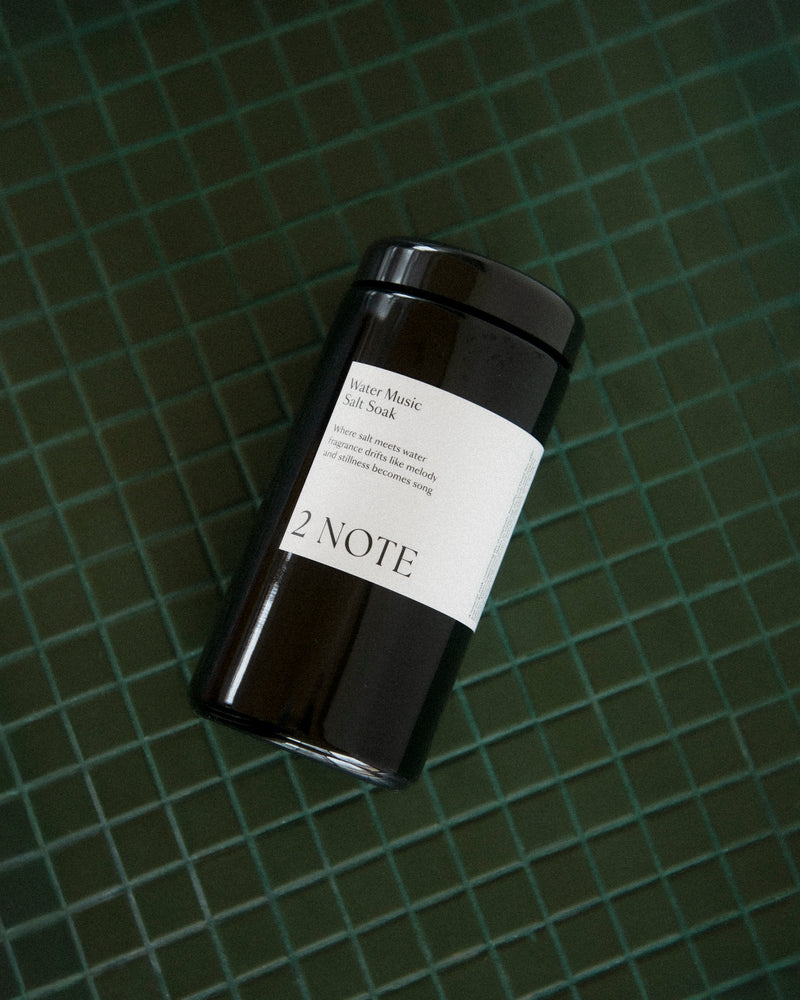 Water Music Salt Soak matte black jar lying on its side on dark green tile — botanical skincare bath soak, moody editorial overhead