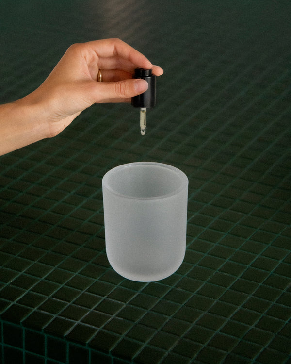 Hand of queer nonbinary model with light olive complexion holding Dwell Drops dropper over frosted white vessel on dark green tile — botanical fragrance drops for home scenting, in use