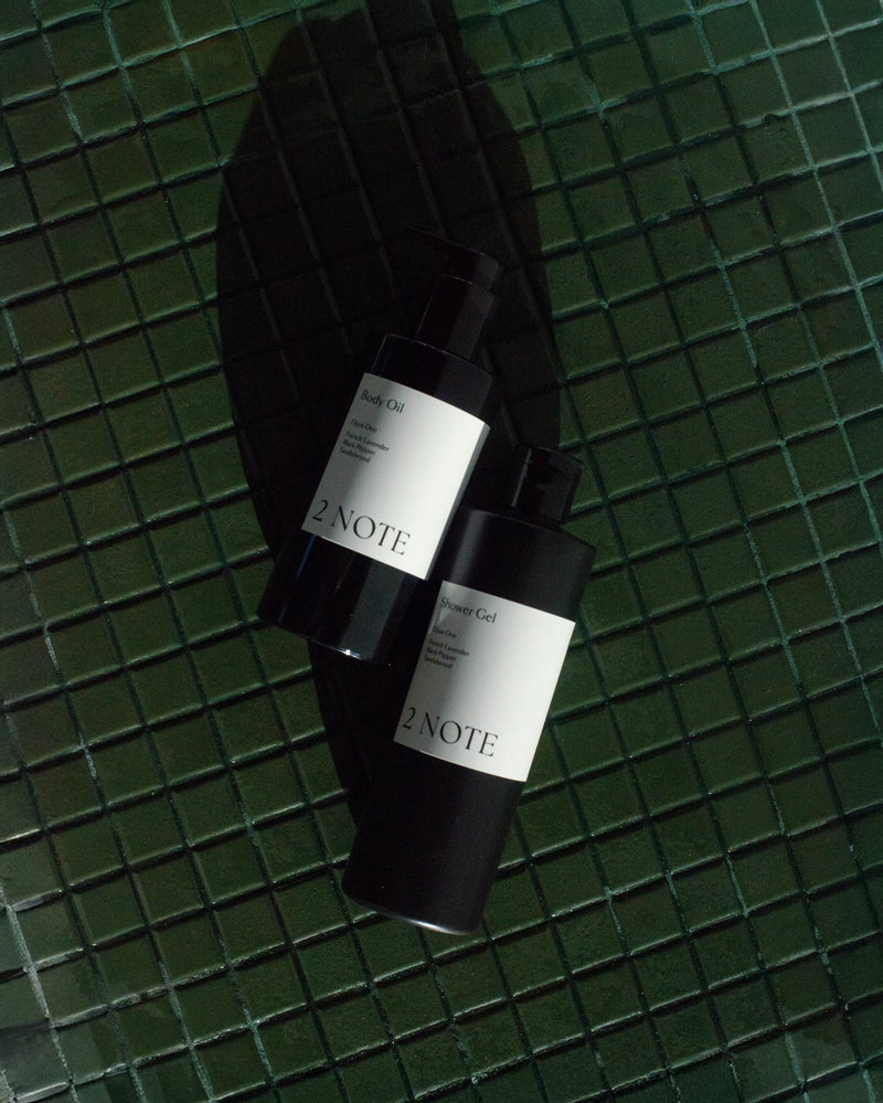 Body Oil and Shower Gel matte black bottles on dark green glazed tile — botanical natural skincare body ritual in dramatic diagonal sunlight