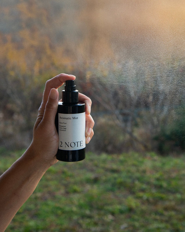 Hand of queer nonbinary model with light olive complexion spraying Aromatic Mist outdoors — botanical fragrance mist in use, visible spray cloud against blurred autumn foliage