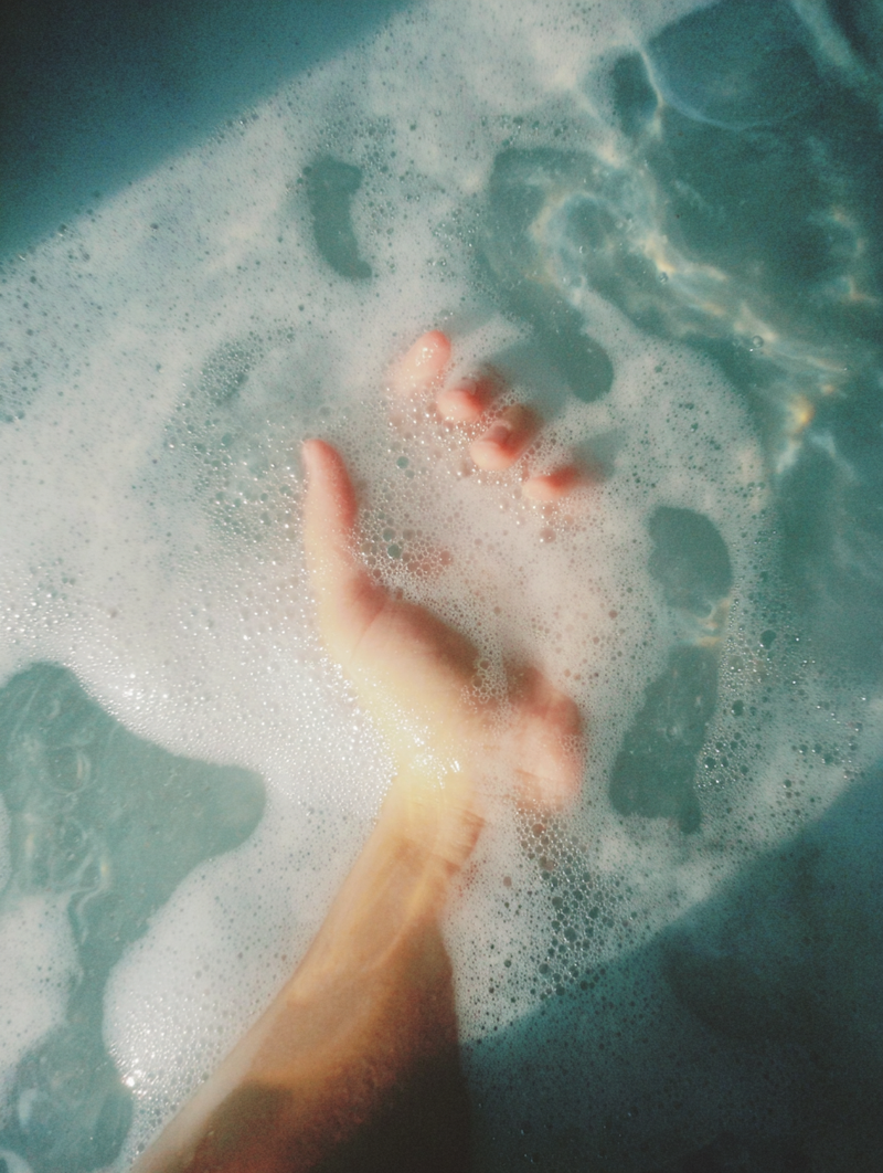Hand of queer nonbinary model with light olive complexion submerged in frothy blue-green bath water — Water Music Salt Soak natural skincare bath soak in use