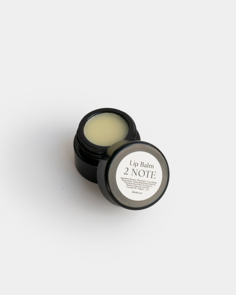 Lip Balm in open small matte black glass jar — botanical natural skincare lip balm showing pale yellow cream texture, overhead on light gray