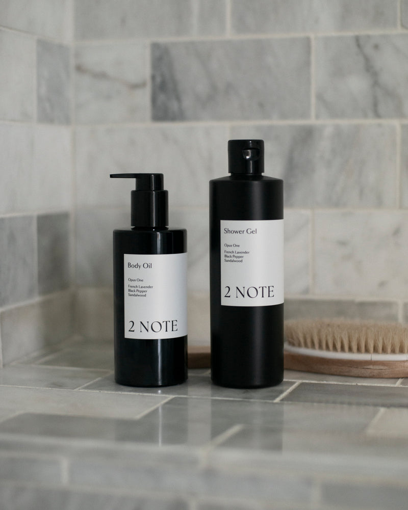 Body Oil and Shower Gel matte black bottles on marble tile shower shelf with natural bristle brush — botanical skincare shower ritual