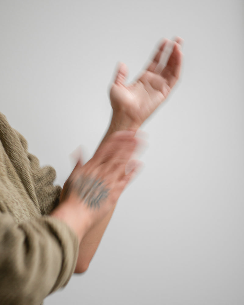 Wrist and forearm with sun tattoo of queer nonbinary model with light olive complexion, applying Luminous Body Oil — botanical skincare brightening body oil in use, motion blur on soft gray
