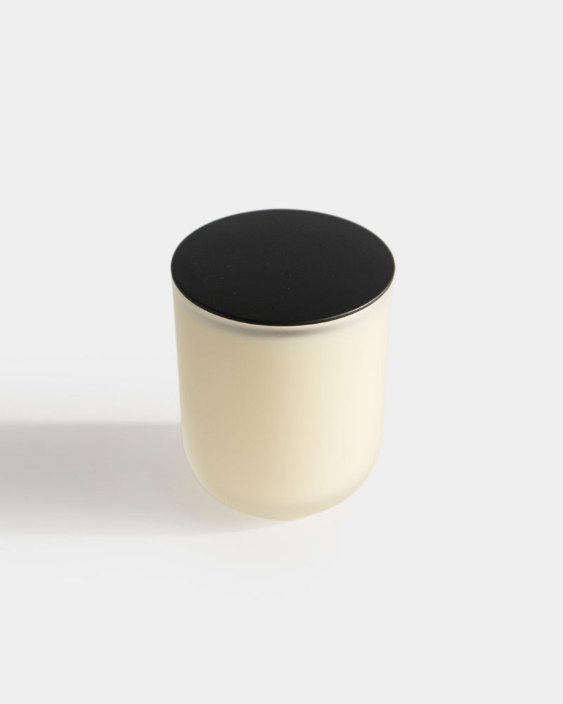 Aromatic Candle in frosted white glass vessel with matte black round lid on top — botanical fragrance candle unlit, front view on light gray