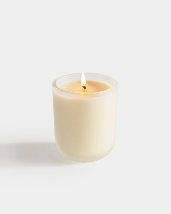 Aromatic Candle lit in frosted white cylindrical glass vessel — hand-poured botanical fragrance candle, cream wax with amber flame, front view on light gray