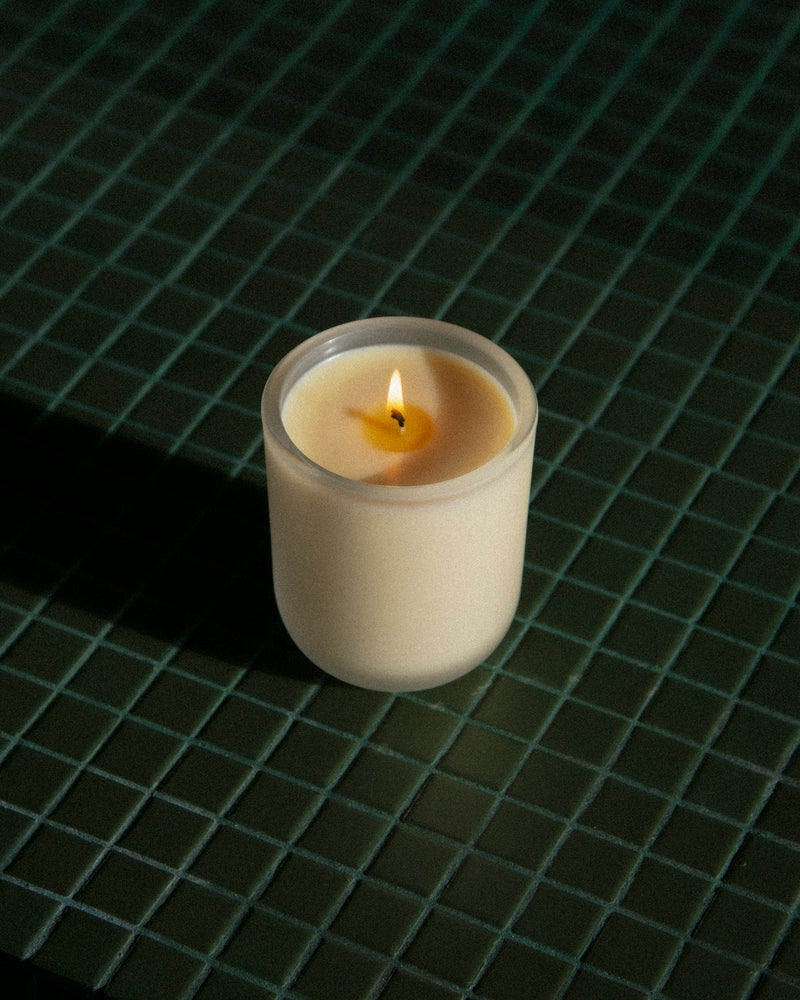 Aromatic Candle lit in frosted white glass vessel on dark green glazed tile — botanical fragrance candle glowing in moody low light