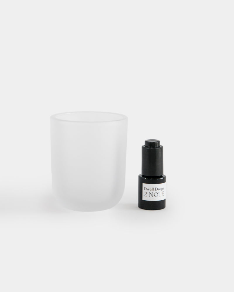 Dwell Drops matte black dropper bottle beside empty frosted white diffuser vessel — botanical home fragrance system, both on light gray