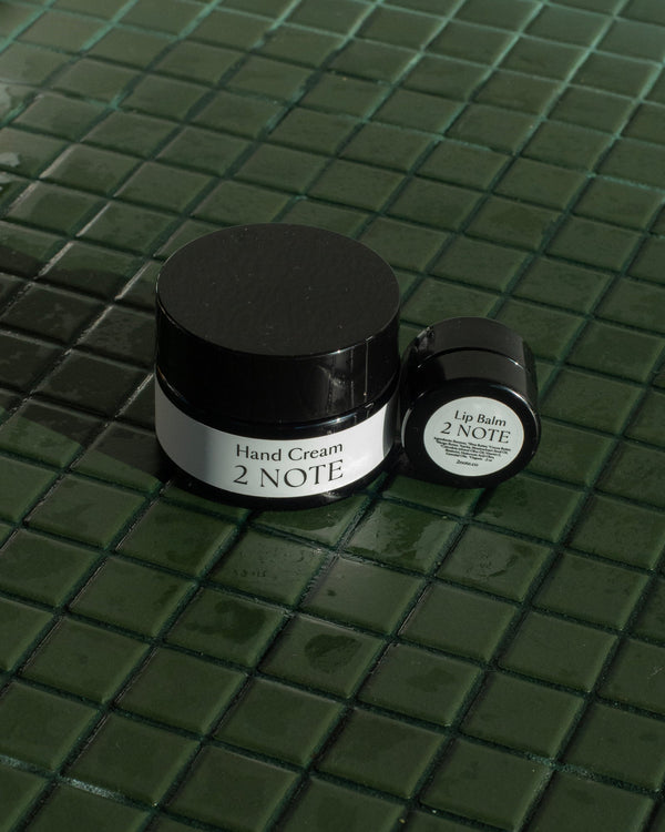Hand Cream and Lip Balm matte black jars on dark green glazed tile — botanical skincare hand and lip care pairing in direct sunlight