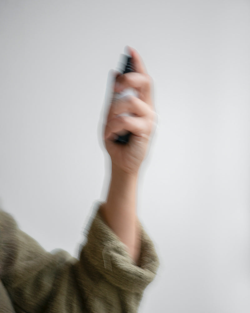 Hand of queer nonbinary model with light olive complexion spraying Resonance Deodorant — botanical natural skincare spray deodorant in motion, motion blur on soft gray