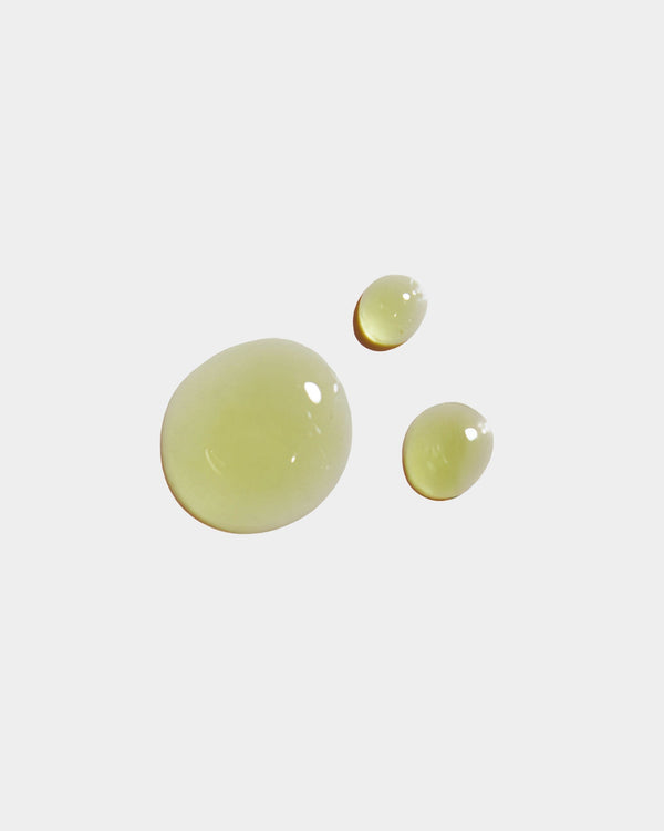 Luminous Body Oil formula swatch — three pale yellow-green botanical oil drops on light gray surface, natural skincare texture